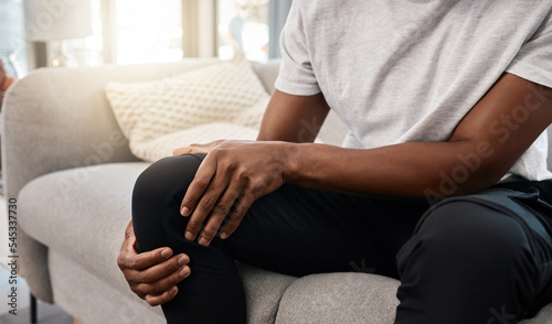 Knee pain, hand of a man and injury while sitting on home living room sofa for rest and relaxation. Ache, painful leg joint and african american man touching his knees for a strain muscle photo