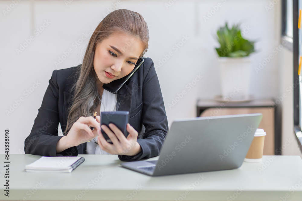 Smiling Asian business woman at work talking on phone and taking notes, sitting at her working place in office, copy space Smiling Asian business woman at work talking on phone and taking notes, sitting at her working place in office, copy space