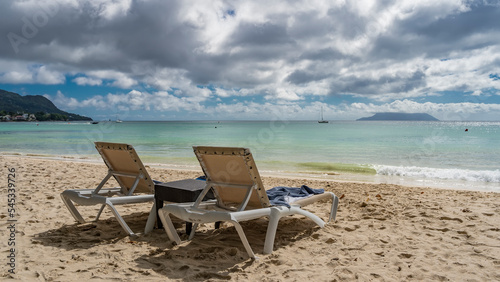 Two sun loungers are on the sandy beach. The waves of the turquoise ocean are foaming. On the horizon, in the clouds, the silhouette of the island is visible. A sunny day. Seychelles. Mahe. 