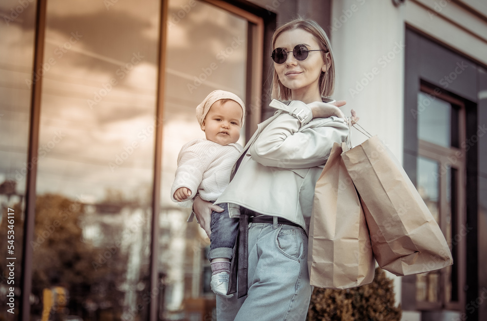 Modern stylish mom with her daughter in her arms and shopping bags in city. Lifestyle