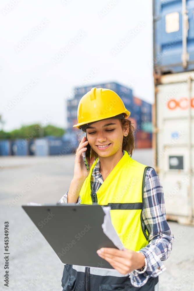 Photo of a young beautiful professional western female brunette engineer cross checking information on the mobile phone about the containers in a shipping containers yard