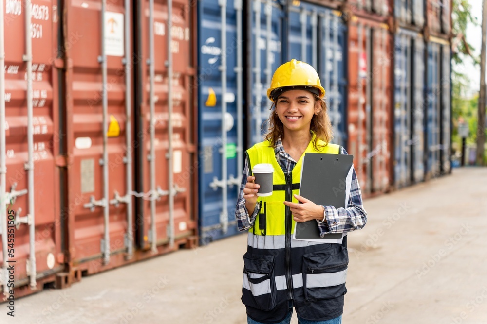 Photo of a young beautiful professional western female brunette engineer holding a coffee cup and document clip board while inspecting containers in a shipping containers yard in the morning