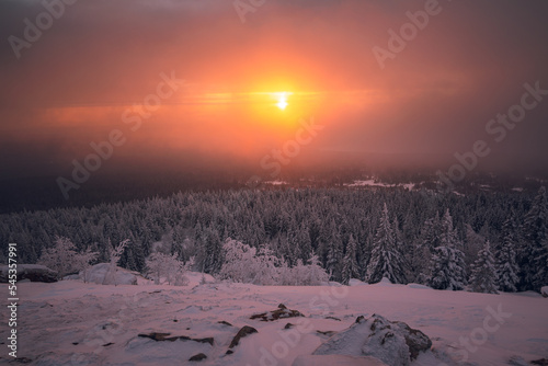 Winter sunrise on the top of the mountain with the sun shines through the clouds above snow-covered trees.