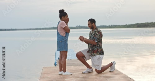 Young cheerful handsome man standing on one knee making proposal of marriage to his beloved woman on dock on lake giving engagement ring. Happy surprised woman got engaged with boyfriend. Love concept
