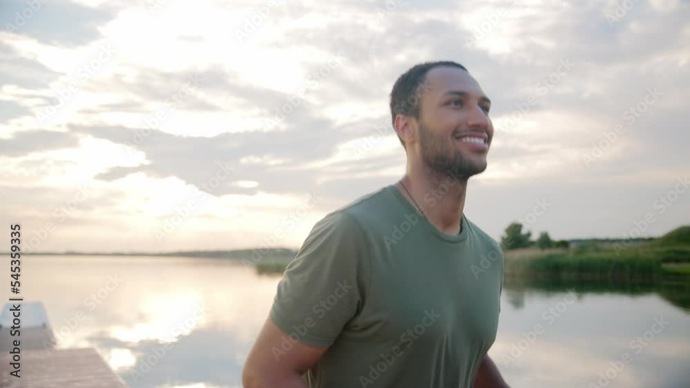 Close up portrait of joyful handsome young sportsman running on nature outdoors in good mood and smiling. Morning workout. African American attractive male athlete runs on dock on lake on sunset Close up portrait of joyful handsome young sportsman running on nature outdoors in good mood and smiling. Morning workout. African American attractive male athlete runs on dock on lake on sunset