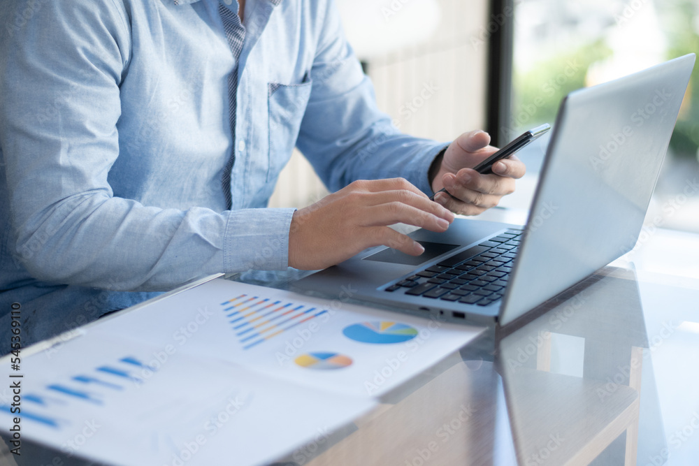 Man in blue shirt typing on laptop and holding phone
