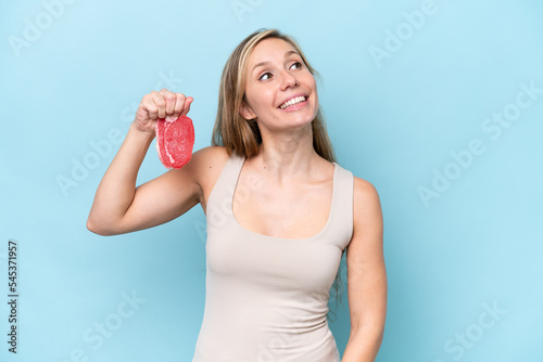 Young blonde woman holding a piece of meat isolated on blue background looking up while smiling