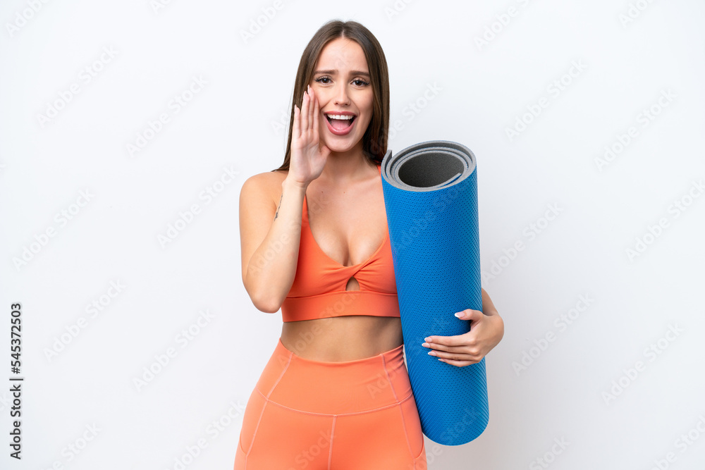 Young beautiful sport woman going to yoga classes while holding a mat isolated on white background shouting with mouth wide open Young beautiful sport woman going to yoga classes while holding a mat isolated on white background shouting with mouth wide open