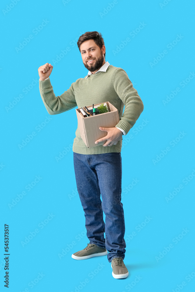 Happy fired young man holding box with personal stuff on blue background Happy fired young man holding box with personal stuff on blue background