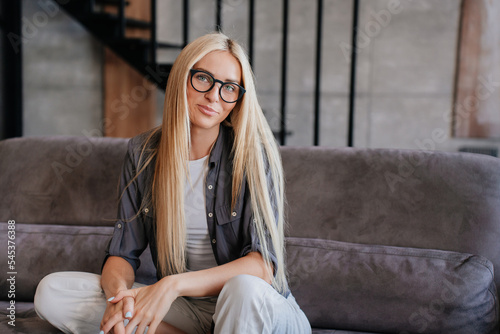 Purposeful blonde caucasian woman in casual sitting  on sofa at home looks at camera confidently. Pretty Swedish girl in glasses with long loose hair spends time on weekend with domestic leisure. © Iona