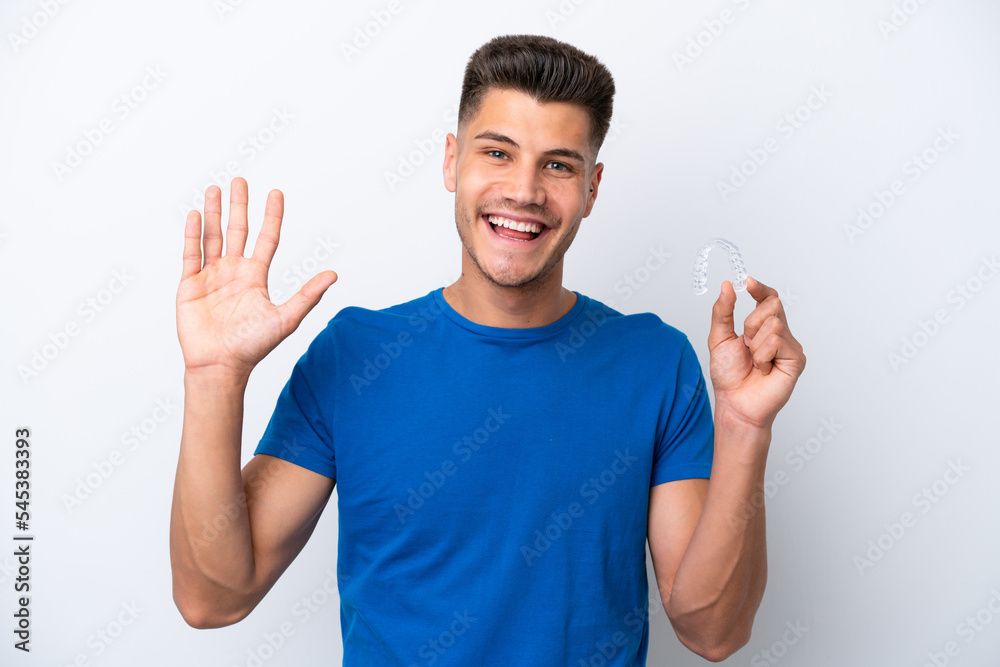 Young caucasian man holding invisible braces isolated on white background saluting with hand with happy expression Young caucasian man holding invisible braces isolated on white background saluting with hand with happy expression
