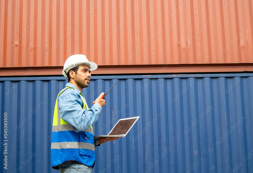 A male inspector in a container shipping company using a laptop and reported through a walkie-talkie A male inspector in a container shipping company using a laptop and reported through a walkie-talkie