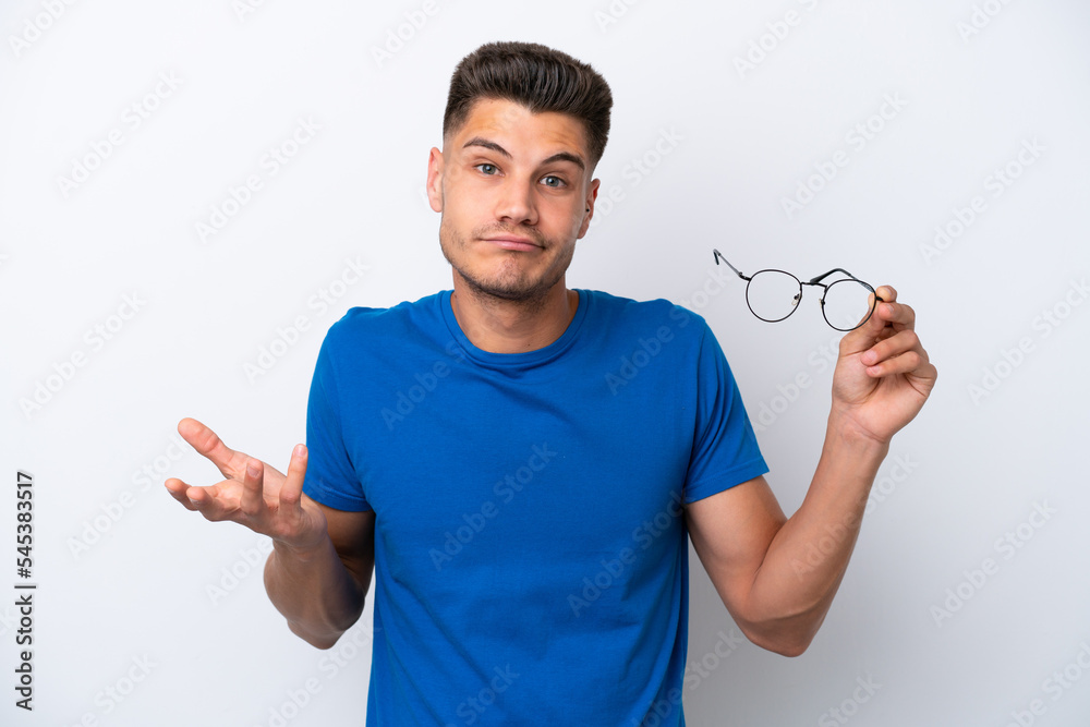 Young caucasian man holding glasses isolated on white background making doubts gesture while lifting the shoulders Young caucasian man holding glasses isolated on white background making doubts gesture while lifting the shoulders