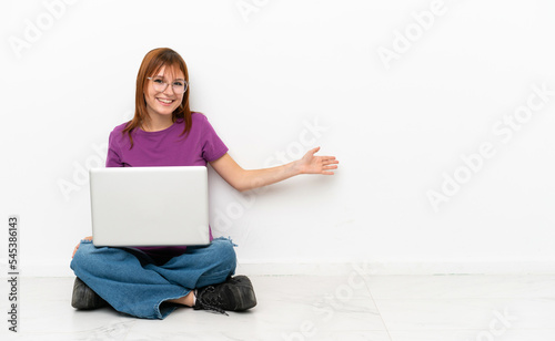 redhead girl with a laptop sitting on the floor extending hands to the side for inviting to come