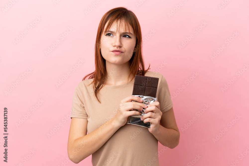 Redhead girl isolated on pink background taking a chocolate tablet and having doubts Redhead girl isolated on pink background taking a chocolate tablet and having doubts