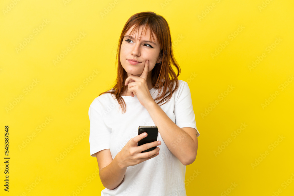 Redhead girl isolated on yellow background using mobile phone and thinking Redhead girl isolated on yellow background using mobile phone and thinking