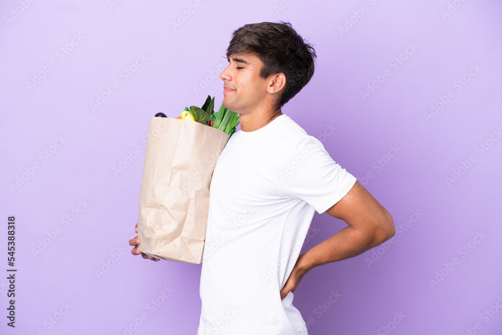 Young man holding a grocery shopping bag isolated on purple background suffering from backache for having made an effort Young man holding a grocery shopping bag isolated on purple background suffering from backache for having made an effort
