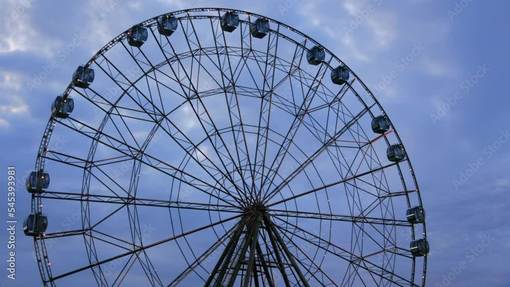 Large and high Ferris wheel with closed cabins spins in the amusement park on the background of the evening blue sky. Entertainment for lovers of extreme entertainment Large and high Ferris wheel with closed cabins spins in the amusement park on the background of the evening blue sky. Entertainment for lovers of extreme entertainment