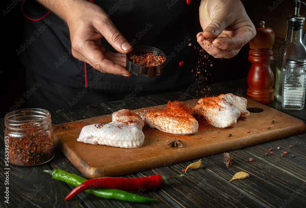 The chef or cook adds fragrant spices to chicken wings. Grilled raw chicken wing in the kitchen of a restaurant or cafe on a kitchen table with vegetables.