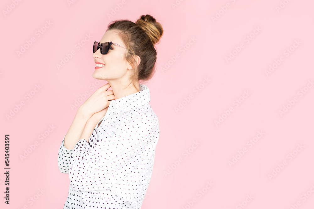 Beautiful girl wearing white shirt and sunglasses posing on pink background in studio. Looking sideways, copy space.