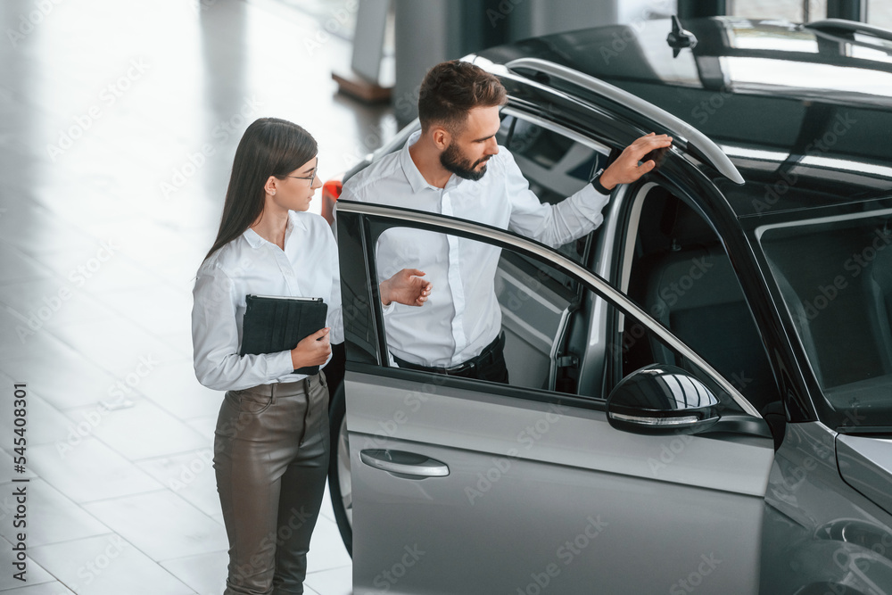 Opening the door. Man with woman in white clothes are in the car dealership together