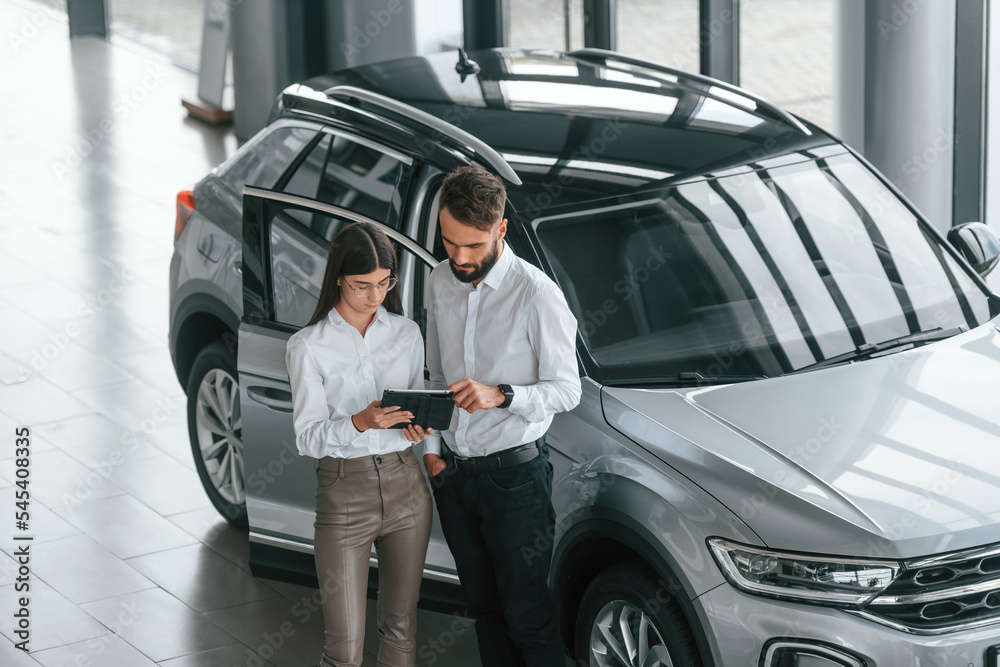 Using digital tablet. Man with woman in white clothes are in the car dealership together