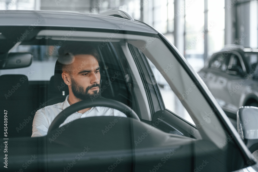 Front view. Sitting inside of automobile. Young man in white clothes is in the car dealership