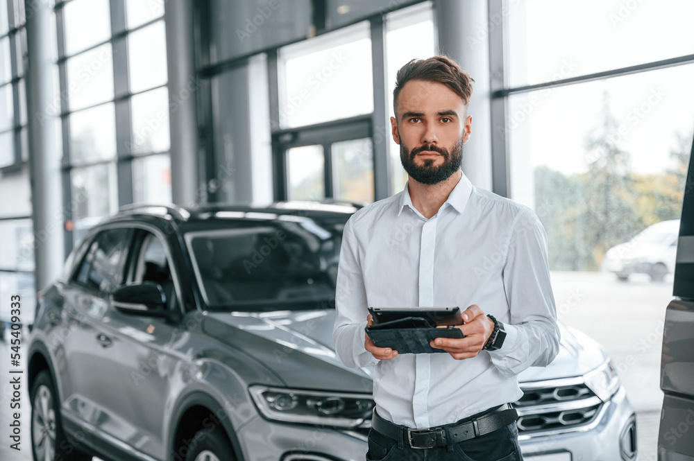 Black graphic tablet in hands. Young man in white clothes is in the car dealership Black graphic tablet in hands. Young man in white clothes is in the car dealership