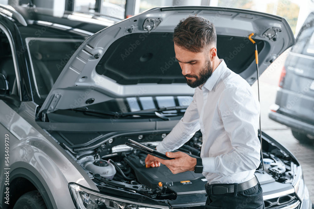Looking under the hood and holding tablet. Young man in white clothes is in the car dealership Looking under the hood and holding tablet. Young man in white clothes is in the car dealership