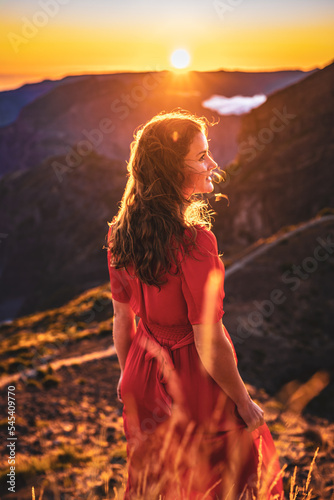 Beautiful brown haired woman in red dress smiling in the sunny evening atmosphere on Pico do Ariero. Verade do Pico Ruivo, Madeira Island, Portugal, Europe.