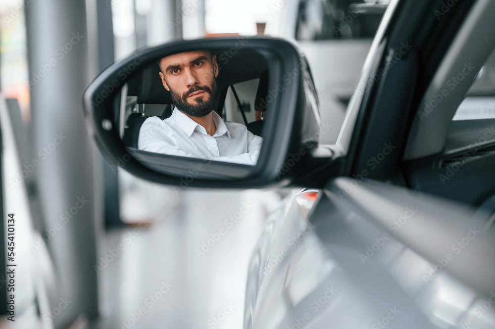 Looking in the side mirror. Young man in white clothes is in the car dealership