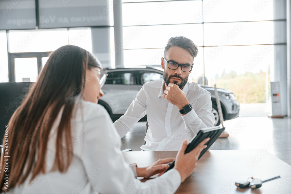 Fototapeta premium Showing information in the tablet. Sitting by the table. Man with woman in white clothes are in the car dealership