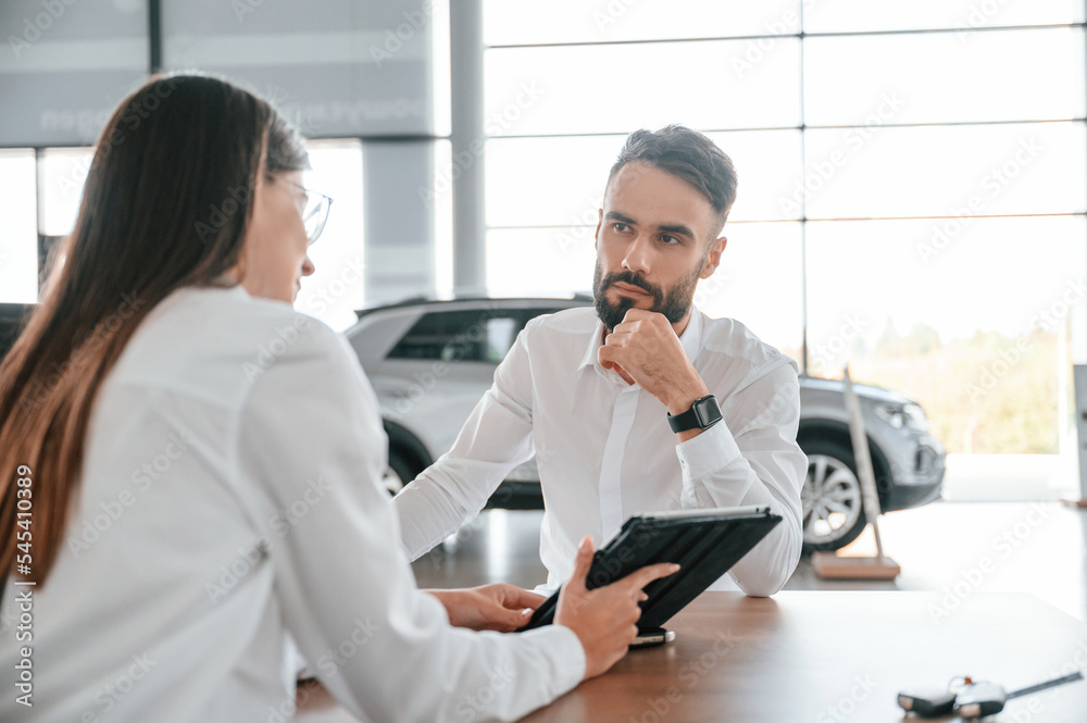 Making a deal. Sitting by the table. Man with woman in white clothes are in the car dealership together Making a deal. Sitting by the table. Man with woman in white clothes are in the car dealership together