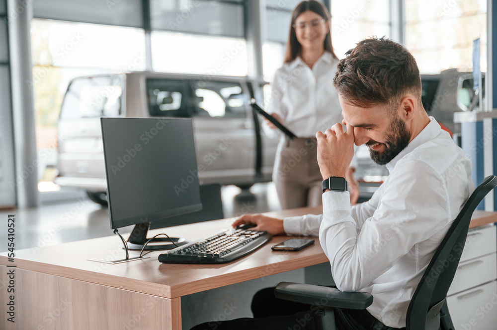 Man is sitting and smiling. Woman in white clothes in the car dealership Man is sitting and smiling. Woman in white clothes in the car dealership