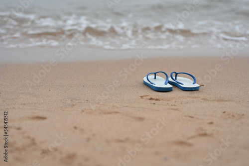 A pair of white flip flops with blue straps rests on sandy beach with a small wave in front of the sea. Sponge shoes were removed and left in wet sand, and there were blurry images of human footprint