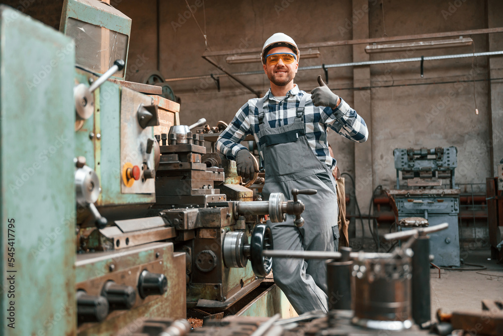 Leaning on the factory machine and showing thumb up. Worker in uniform is indoors