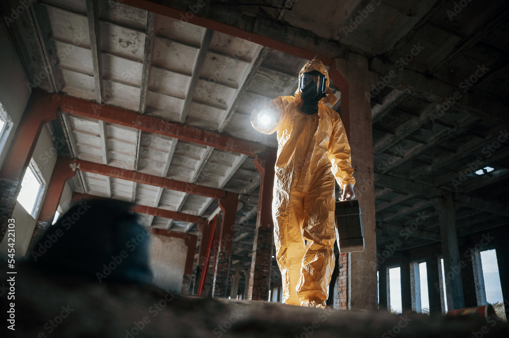 Using the flashlight. Man dressed in chemical protection suit in the ruins of the post apocalyptic building