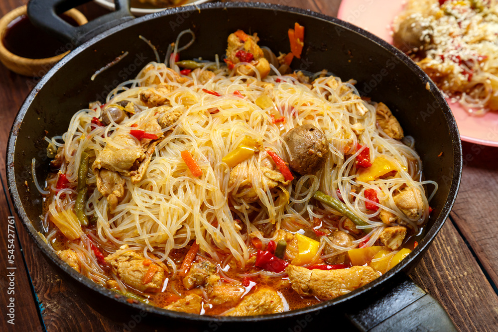 Funchoza with chicken and vegetables in teriyaki and oyster sauce in a frying pan on a wooden table with food. Funchoza with chicken and vegetables in teriyaki and oyster sauce in a frying pan on a wooden table with food.