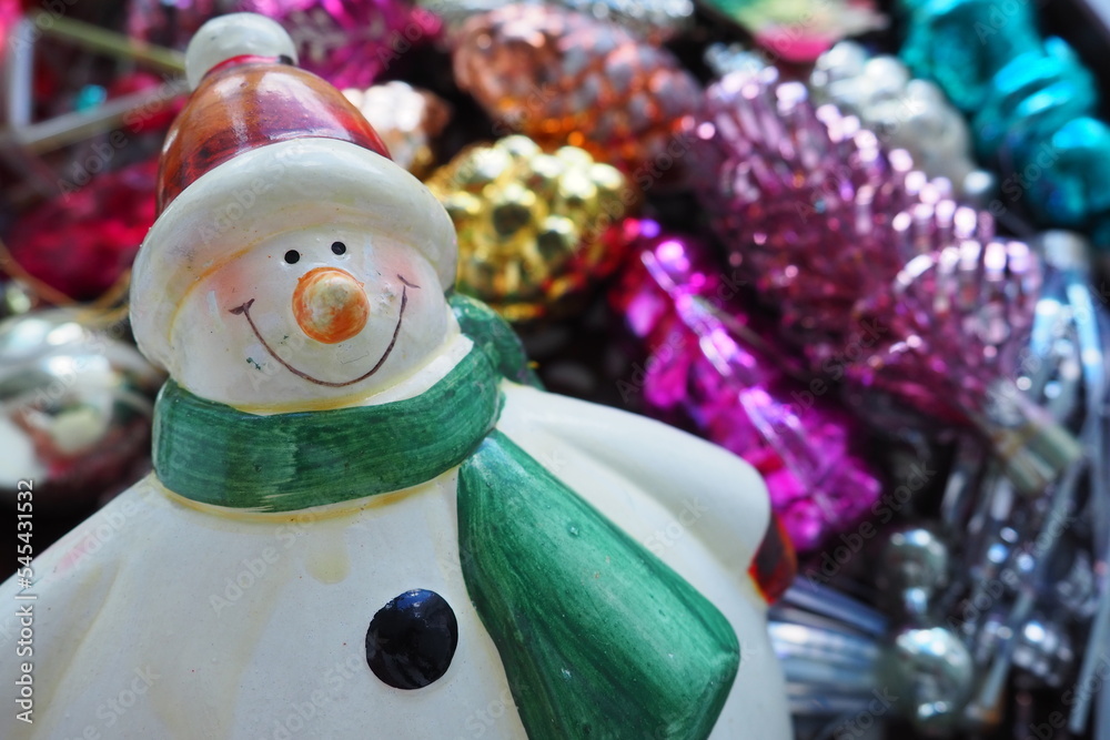 Cheerful smiling ceramic snowman in a green scarf and winter hat. New Year and Christmas decorations, tinsel. Multi-colored balls in the form of cones. Festive interior decor. New Year or Christmas