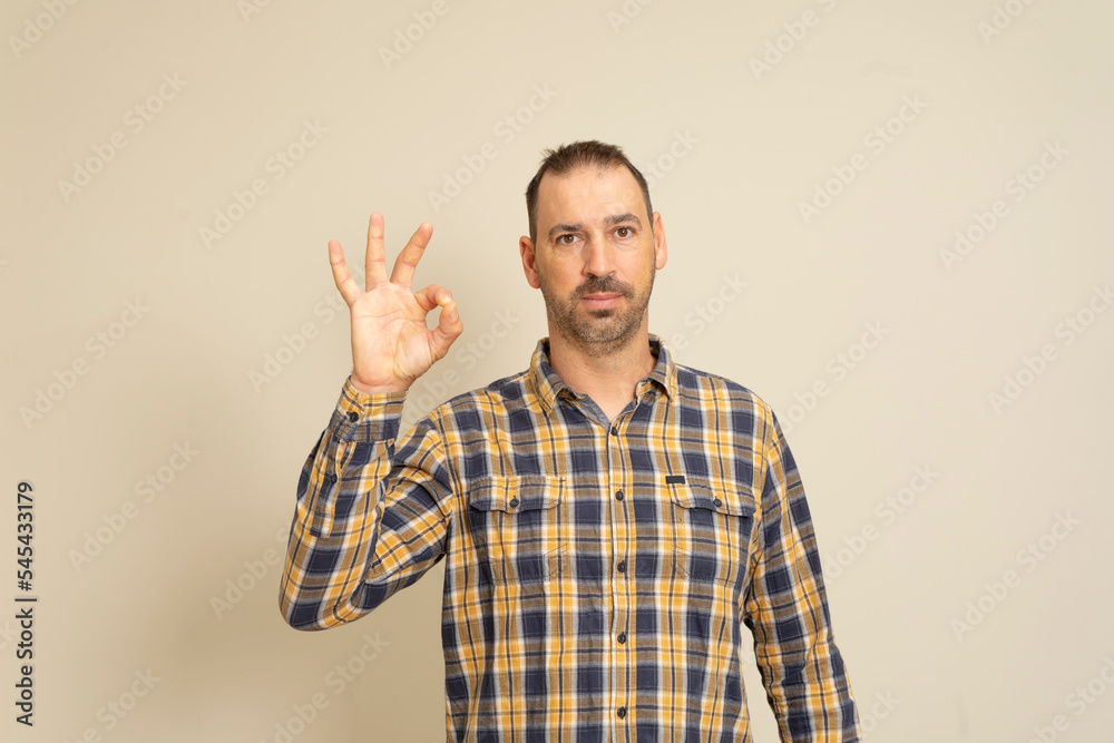 Handsome funny smiling man in checkered shirt posing isolated on beige wall background, studio portrait. People candid emotions lifestyle concept. Mock up copy space. Showing OK gesture.
