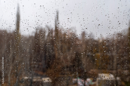 Textured surface of drops of autumn cold rain on window glass against blurred background of an empty dark city park and gray sky. Rainy weather.