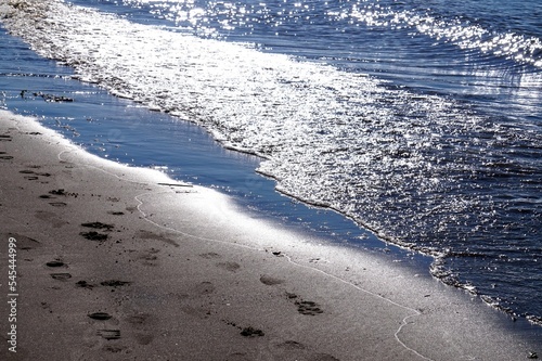 Wasserwellen am Strand bildf  llend verlaufen quer durch das Bild Sonne glitzert auf dem Wasser im Vordergrund Ufer feuchter Sand mit Fu  spuren