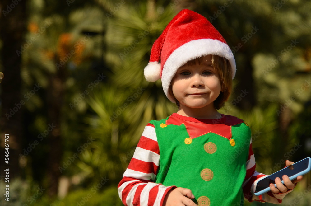 A cute positive little boy in a Santa Claus hat is talking on a smart phone. Concept: Christmas on the beach, tropical resort, child asking for gifts, holiday expectations, family trip. A cute positive little boy in a Santa Claus hat is talking on a smart phone. Concept: Christmas on the beach, tropical resort, child asking for gifts, holiday expectations, family trip.
