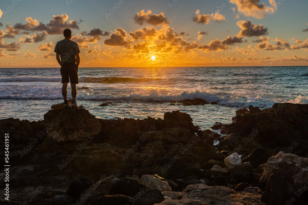 Man, person, silhouette, standing on basalt rocks from an ancient lava flow by the edge of the ocean under cloudy sky watching the sunrise, Poipu, Kauai, Hawaii