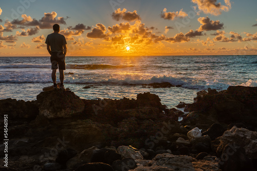 Man, person, silhouette, standing on basalt rocks from an ancient lava flow by the edge of the ocean under cloudy sky watching the sunrise, Poipu, Kauai, Hawaii