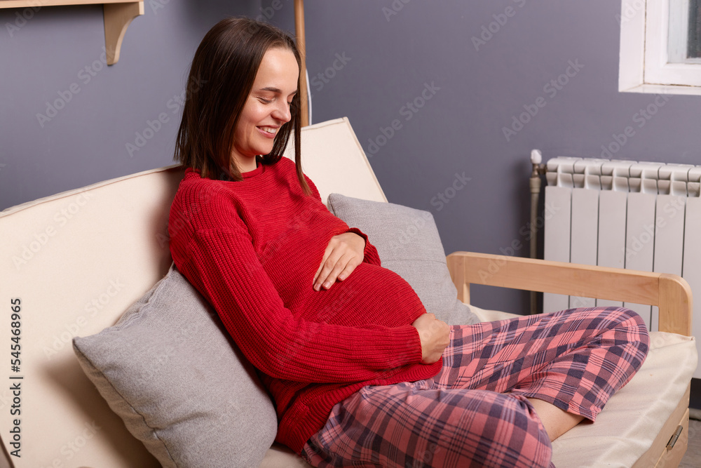 Image of young adult pregnant smiling woman wearing red jumper and pajama pants sitting on sofa in home interior, holding hands on her belly, waiting her baby. Image of young adult pregnant smiling woman wearing red jumper and pajama pants sitting on sofa in home interior, holding hands on her belly, waiting her baby.