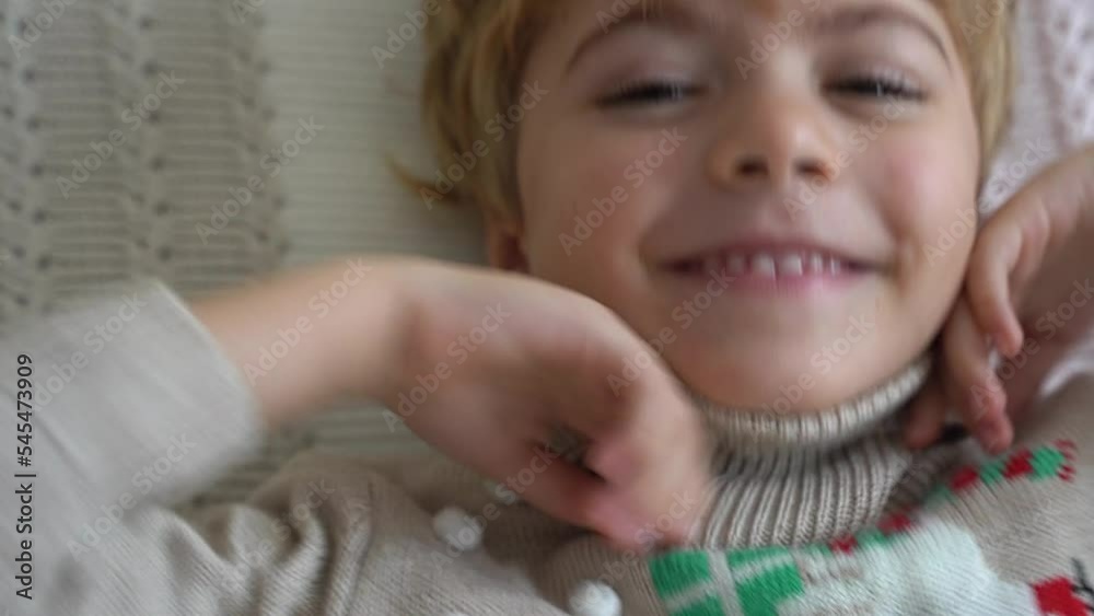 Cute new year child lying on bed looking at camera, smiling. Christmas morning, happiness after getting gifts. Wear Scandinavian sweater with reindeer. Top view. Lifestyle.