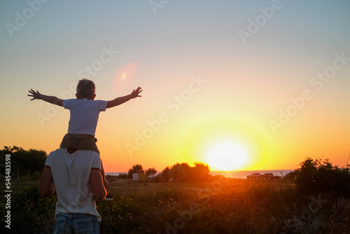 Dad carries a little son on his shoulders walking across the field against the backdrop of a sunset in the sky, back view