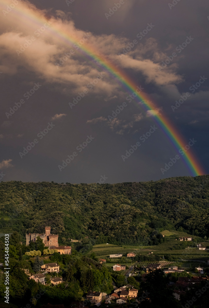 Beautiful rainbow over a medieval rural village with a castle upon a green hill at twilight. Italy, Piedmont.