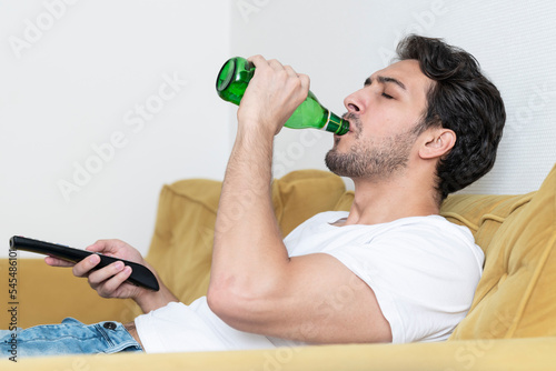 Handsome man lying on the couch and watching tv and drinking beer 